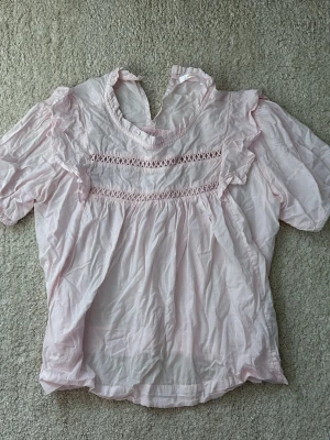 Ljusrosa blus med volangdetaljer - Söt ljusrosa blus med korta puffärmar och volangdetaljer vid axlarna. Blusen har rund halsringning med liten volangkant och dekorativa spetsband framtill. Tillverkad i ett tunt, mjukt tyg som ger en romantisk känsla.