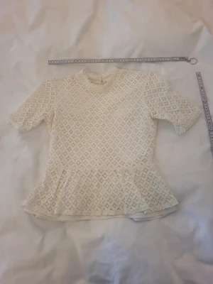 Vit spetsblus med volangkant - Söt vit blus i spets med korta ärmar och volangkant nedtill. Blusen har ett diskret mönster, hög hals och knapp i nacken. Perfekt för dig som gillar romantisk och feminin stil.