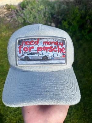 Ljusgrå keps från Revenite - Säljer en ljusgrå keps från Revenite med mesh på baksidan och justerbar snapback. Framtill finns ett stort tygmärke med texten 'need money for porsche' och en bild på en Porsche. Kepsen har en modern streetwear-vibe och är tillverkad i mjukt material. Limiterad finns bara i en antal upplaga