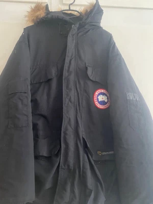 Svart dunjacka från Canada Goose - Säljer en svart dunjacka från Canada Goose med klassisk patch på bröstet och flera praktiska fickor. Jackan har huva med fuskpäls och är perfekt för kalla vinterdagar. Oversized passform och robust dragkedja framtill.