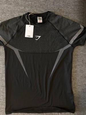 Gymshark onyx 5.0 - Snygg svart tränings-tshirt från Gymshark med gråa detaljer och mönstrade partier över axlar och bröst. Tillverkad i ett lätt och snabbtorkande material som passar perfekt för gymmet eller löpning. Sportig design med Gymshark-logga på bröstet.