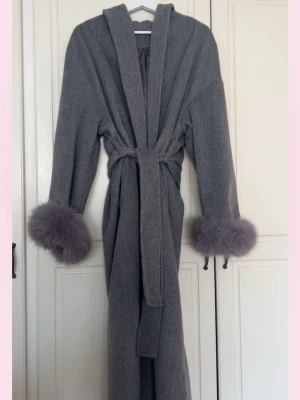Grå lång kappa med päls arm - 💞Stilren grå lång kappa med skärp i midjan och breda ärmar dekorerade med fluffiga päls arm i matchande grå ton. Kappan har en klassisk krage och rak passform, perfekt för dig som vill ha en trendig och elegant look.💞
