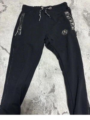 Svarta mjukisbyxor från Philipp Plein - Säljer ett par svarta mjukisbyxor från Philipp Plein med coola ormmönstrade detaljer vid fickorna och logopatch på benet. Byxorna har dragsko i midjan och smal passform. Perfekta för dig som gillar streetwear och vill sticka ut.