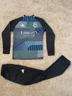 Real Madrid träningsset Adidas - Snyggt träningsset från Adidas och Real Madrid. Tröjan har lång ärm, svart bas med mönstrade paneler i blått, grönt och grått samt klubbmärke och sponsortryck. Byxorna är svarta med Adidas-logga. Perfekt för fotbollsträning eller gymmet.