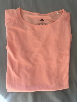 Ljusrosa Adidas träningstopp XS - Ljusrosa träningstopp från Adidas med rund hals och korta ärmar. Tillverkad i ett ribbat funktionsmaterial som andas, perfekt för träning. Adidas-logga i silver nedtill på framsidan.