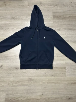 Mörkblå hoodie från Polo Ralph Lauren - Snygg mörkblå hoodie från Polo Ralph Lauren med dragkedja framtill och klassisk logga broderad på bröstet. Hoodien har justerbar huva med snören och två stora fickor framtill. Tillverkad i mjukt bomullsmaterial för extra komfort.