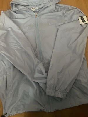 Ljusblå vindjacka från Risky - Ljusblå vindjacka från Risky med huva och dragkedja framtill. Jackan har elastiska muddar vid ärmsluten, sidofickor och en patch med motiv på ena ärmen. Tillverkad i lätt och vindtätt material, perfekt för blåsiga dagar.