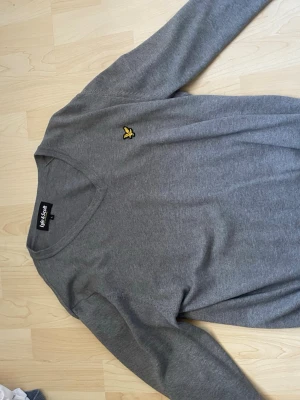 Grå tröja från Lyle & Scott - Snygg grå tröja från Lyle & Scott med klassisk gul logga på bröstet. Tröjan har rund halsringning och långa ärmar. Tillverkad i mjukt material som känns skönt mot huden. Perfekt för en clean och stilren look.