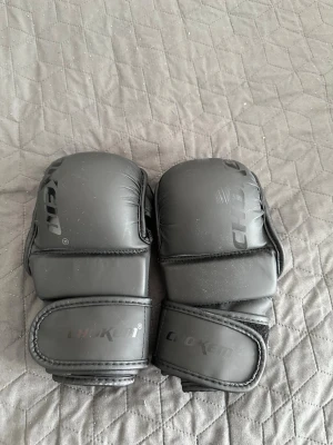 Mma Handskar  - Säljer ett par svarta MMA-handskar från CHOKEN med öppen design för fingrarna och vadderad ovansida. Handskarna har bred kardborrestängning runt handleden för extra stöd och är tillverkade i konstläder. Perfekta för kampsportsträning. 