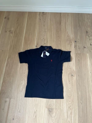 Marinblå piké från Polo Ralph Lauren - Snygg marinblå pikétröja från Polo Ralph Lauren med klassisk krage och korta ärmar. Tröjan har den ikoniska röda logotypen broderad på bröstet och är tillverkad i mjuk bomull för en bekväm känsla.