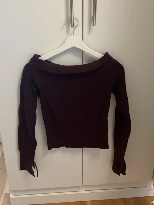 Vinröd off shoulder stickad tröja - Säljer en vinröd stickad tröja med off shoulder-ringning och långa ärmar. Tröjan har ribbstickad kant upptill och fina flätstickade detaljer längs ärmarna. Ärmsluten har dekorativa knytband som ger en extra snygg touch.