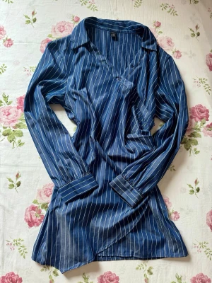 Blå randig skjorta med v-ringning - Snygg blå skjorta med vita ränder och v-ringning. Skjortan har långa ärmar med breda manschetter och klassisk krage. Materialet känns mjukt och har ett lätt glansigt utseende, perfekt för en trendig look.