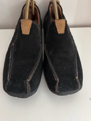 Svarta mockaloafers med kontrastsömmar - Säljer ett par svarta loafers i mocka med bruna kontrastsömmar och detaljer. Skorna har en fyrkantig tå och dekorativa nitar på sidan. Insidan är fodrad i ljust material och de har en låg sula. Perfekta för en avslappnad men ändå stilren look.