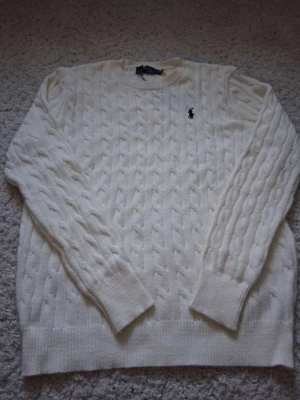 Vit kabelstickad tröja Polo Ralph Lauren - Kabelstickad vit tröja från Polo Ralph Lauren med klassisk rund halsringning och svart broderad logga på bröstet. Tröjan har långa ärmar och ribbade muddar vid ärmslut och nederkant. Perfekt för dig som gillar stilrena och tidlösa plagg. Passar mer som S.