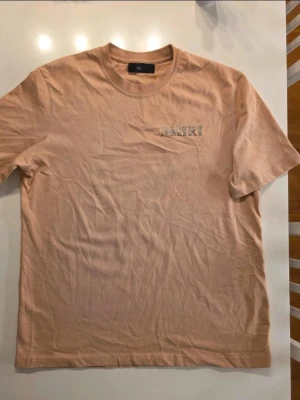 Beige AMIRI t-shirt i bomull - Snygg beige t-shirt från AMIRI i 100% bomull. Stor AMIRI-logga på ryggen och mindre logga på bröstet. Klassisk passform med rund hals och korta ärmar. Metallbricka med AMIRI vid nederkanten ger en exklusiv touch. Jättesnygg oversize t-shirt  Bröstmått: ca 55 cm Längdmått: ca 72 cm