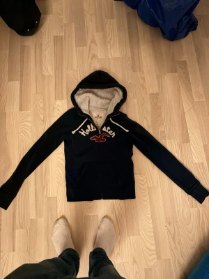 Marinblå hoodie från Hollister - As fet marinblå hoodie från Hollister med dragkedja. Fodrad huva med vitt teddyfoder och vita dragsnören. Perfekt för dig som gillar klassisk college-stil och vill ha något riktigt bekvämt. Jättebra skick och pris kan diskuteras 