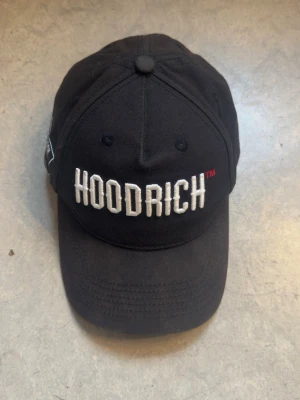Hoodrich cap - Säljer en svart keps från Hoodrich med böjd skärm och broderad vit logga framtill. Kepsen har en klassisk sexpanelsdesign och en liten etikett på sidan med texten 'HOOD-TECH'. Tillverkad i bomullstyg.