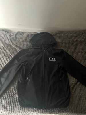 Svart EA7 vindjacka med huva - Snygg svart vindjacka från EA7 Emporio Armani med huva och dragkedja framtill. Jackan har två fickor med dragkedja och logga på bröstet. Tillverkad i lätt och tåligt material, perfekt för blåsiga dagar.