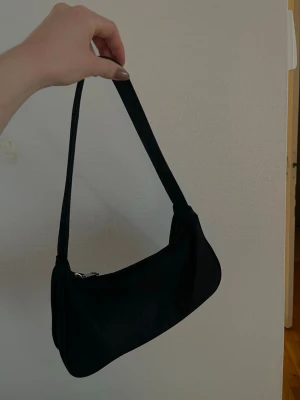 Svart axelväska i minimalistisk stil - En svart axelväska med enkel och stilren design. Väskan har en mjuk, något böjd form och ett brett axelband. Stängs med dragkedja upptill och är tillverkad i ett slätt tygmaterial. Från Asos, bra skick! 