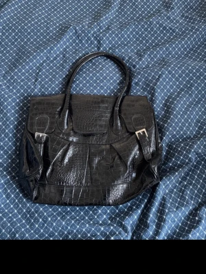 Svart handväska i krokodilpräglat skinn - Snygg svart handväska i fejkat krokodilskinn med två handtag och silverfärgade spännen. Väskan har en klassisk fyrkantig form och lock med spänndetaljer. Perfekt för dig som gillar en trendig och stilren look. Hittar inte vart den är ifrån säljer även på vinted om man föredrar de