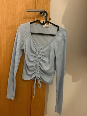 Ljusblå ribbad topp med snörning - Trendig ljusblå långärmad topp från Monki med ribbad struktur och snygg snörning framtill. V-ringad och croppad modell som ger en modern look. Perfekt att matcha med höga jeans eller kjol.