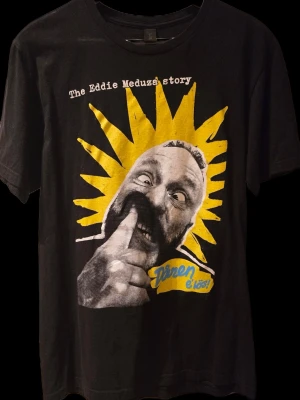 Svart Eddie Meduza t-shirt - Cool svart t-shirt med tryck från 'The Eddie Meduza story'. Framsidan har ett stort grafiskt motiv i gult, vitt och blått med text och illustration. Klassisk passform och rund halsringning. Perfekt för dig som gillar unik och retro stil.