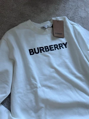 Vit sweatshirt från Burberry - Säljer en stilren vit sweatshirt från Burberry med svart logotyp tryckt framtill. Tröjan har rund halsringning och långa ärmar. Tillverkad i mjuk bomull för en bekväm känsla. Perfekt för dig som gillar klassisk och enkel design.