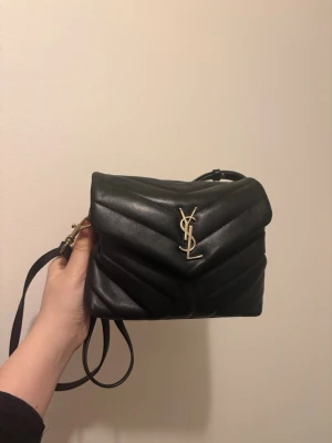 Svart YSL handväska i läder - Säljer en svart handväska från Yves Saint Laurent i mjukt läder med quiltat mönster och guldfärgat YSL-logo på framsidan. Väskan har axelrem och en stilren, modern design som passar till många olika looks. Ingen kartong eller kvitto! Den har gått sönder se bild! Går ha som clutch eller använda ändå såklart de bilder