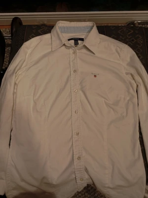 Vit skjorta från Gant  - Klassisk vit skjorta från Gant med lång ärm och knappar framtill. Skjortan har en liten broderad logga på bröstet och en stilren krage. Tillverkad i bomull för en skön känsla och snygg passform. Perfekt för bröllop eller andra fina kalas. Nypris runt 1000 kr. Använd fåtal gånger.