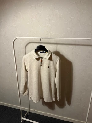 Beige långärmad polotopp från Polo Ralph Lauren - Klassisk beige långärmad polotopp från Polo Ralph Lauren med krage och half zip-dragkedja. Diskret broderad logga på bröstet och mjukt material som känns skönt mot huden. Perfekt till jeans eller chinos för en clean look.