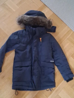 Mörkblå parkas från Didriksons stoelek 110 barn - Snygg mörkblå parkas från Didriksons med avtagbar huva och fuskpäls. Jackan har stora fickor framtill, dragkedja med vindslå och orange detalj vid dragkedjan. Fodrad med mjuk teddy på insidan och är helt vattentät. Perfekt för kalla dagar