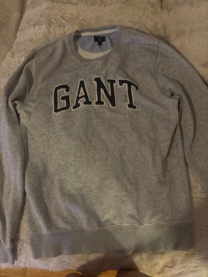 Grå sweatshirt från GANT - Snygg grå sweatshirt från GANT med klassisk rund halsringning och stora svarta bokstäver framtill. Tröjan har ribbade muddar vid ärmslut och nederkant. Perfekt för en avslappnad och stilren look.