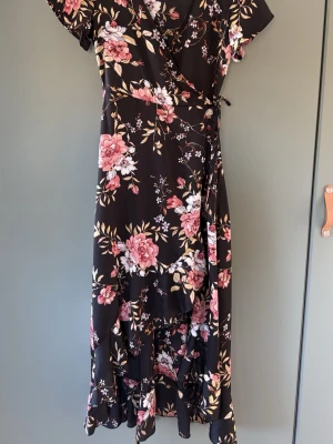 Blommig omlottklänning Chiara Forti - Svart omlottklänning från Chiara Forti med blommigt mönster i rosa, vitt och beige. Klänningen har volangdetaljer, korta ärmar och knyt i midjan. Perfekt för dig som gillar romantisk stil och vill ha en klänning med flowy känsla.