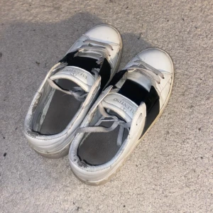 Valentino Garavani vita sneakers - Säljer ett par snygga Valentino Garavani sneakers i vitt skinn med bred svart detalj över sidan. Skorna har rund tå, platt sula och klassisk snörning. Lite slitna men fortfarande väldigt fina (nypris 7800kr) 