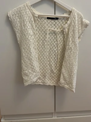 Vit virkad väst från 157 - Säljer en söt vit virkad väst från 157 med knytband fram. Västen har ett luftigt hålmönster och korta ärmar, perfekt att slänga över en topp för en chill look. Materialet känns mjukt och lätt, och den har fina virkade detaljer längs kanterna.