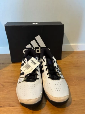 Adidas tennis skor  - Snygga vita tennis skor från Adidas med klassiska svarta ränder på sidorna och svart sula. Skorna har perforerad tå för ventilation, svart innersula och snörning. Perfekt för dig som gillar sportig och stilren look. Kommer med originalkartong. Passar även storlek 44