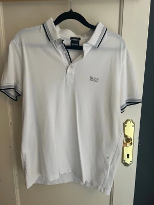 Vit pikétröja från BOSS - Klassisk vit pikétröja från BOSS med diskret logga på bröstet och kontrasterande mörka ränder på krage och ärmslut. Tröjan har korta ärmar och knäppning framtill. Perfekt för en clean och stilren look. På sista bilden ser du att tröjan har en liten fläck. Passar om du är runt 170. Storlek: Small