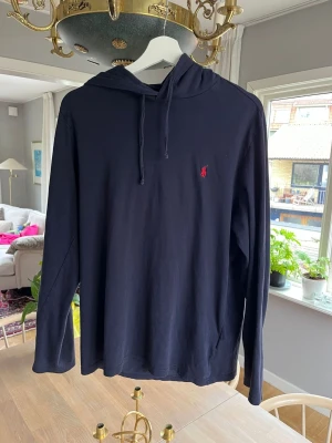 Mörkblå hoodie från Polo Ralph Lauren - Snygg mörkblå hoodie från Polo Ralph Lauren med klassisk huva och dragsko. Liten röd logga broderad på bröstet. Tillverkad i mjukt bomullsmaterial och har långa ärmar. Perfekt för en avslappnad och stilren look.