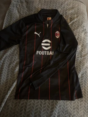 AC Milan träningströja från Puma - Snygg svart långärmad träningströja från Puma med AC Milan-logga på bröstet och röda tunna ränder. Halvlång dragkedja framtill och vit Puma-logga. Perfekt för fotbollsträning eller som supporterplagg.