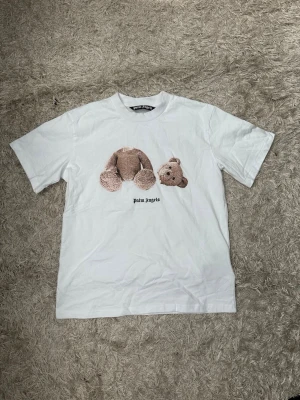 Vit Palm Angels t-shirt med björntryck - Säljer en vit t-shirt från Palm Angels med ikoniskt björntryck framtill och stor logga i svart på ryggen. T-shirten har normal passform och är kortärmad, tillverkad i mjuk bomull. Perfekt för dig som gillar streetwear och vill sticka ut. Storlek: S/M