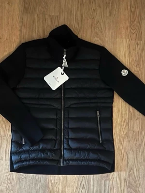 Moncler Cardigan  - Svart dunjacka från Moncler med quiltad front, två dragkedjefickor och hög krage. Jackan har Moncler-logga på ärmen och stilren design. Materialmix med dun och textil ger både värme och en modern look. Perfekt för dig som gillar exklusiva streetwear-plagg.