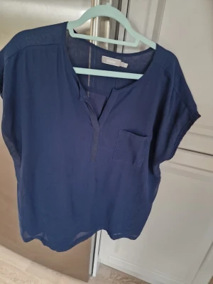 Mörkblå blus från Fransa - Snygg mörkblå blus från Fransa med avslappnad passform och korta ärmar. Toppen har en v-ringad hals och en bröstficka som detalj. Perfekt till jeans eller kjol för en enkel men stilren look.