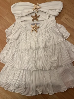 Vit axelbandslös volangklänning - Supersöt vit axelbandslös klänning med flera lager volanger och tre dekorativa sjöstjärnor framtill. Klänningen har plisserad kjol och ger en riktigt somrig känsla. Perfekt för dig som vill sticka ut med en unik detalj!