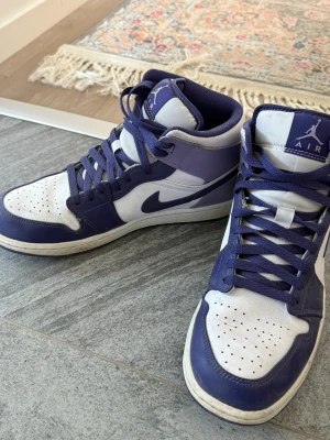 Nike Air Jordan 1 High vit/lila - Säljer ett par Nike Air Jordan 1 High sneakers i vitt och lila. Klassisk siluett med högt skaft, perforerad tåbox och ikoniska swoosh-loggan på sidan. Ovandelen är i läder och skorna har matchande lila snören och detaljer.