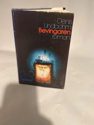 Bevingaren av Dénis Lindbohm - Science fiction-roman med hård pärm och skyddsomslag. Omslaget är svart med futuristiskt motiv i blått och orange samt titeln i röd och blå text. Boken är skriven av Dénis Lindbohm och utgiven av Bokförlaget.