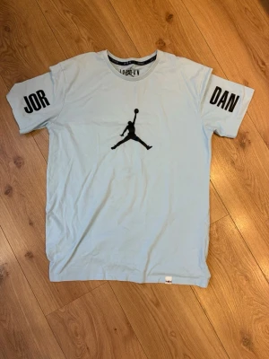 Jordan t-shirt  - Säljer en ljusblå t-shirt från Jordan med det klassiska Jumpman-trycket i svart på bröstet och texten 'JOR' och 'DAN' på ärmarna. T-shirten är i 100% bomull och har en avslappnad passform. Perfekt för dig som gillar streetwear och sportig stil.