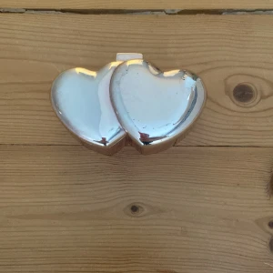 Smyckesask i form av dubbla hjärtan - Säljer en söt smyckesask i blankt silverfärgat metallmaterial, formad som två sammanlänkade hjärtan. Insidan är klädd i svart tyg för att skydda dina smycken. Perfekt för att förvara ringar, örhängen eller små accessoarer.