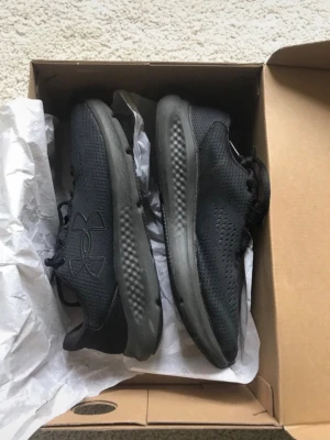 Svarta Under Armour sneakers - Snygga helsvarta sneakers från Under Armour med ventilerande mesh-ovandel och diskret logga på sidan. Skorna har en låg profil och en bekväm, stötdämpande sula som passar perfekt till en aktiv livsstil. Klassisk snörning och sportig look.