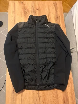 Svart vindjacka från karhu - Snygg svart vindjacka från karhu med quiltad front och rygg, hög krage och dragkedja hela vägen. Jackan har långärmade släta ärmar och reflekterande detaljer på ryggen. Perfekt för kyliga dagar och outdoor-aktiviteter.