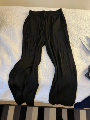 Svarta vida satinbyxor med dragsko - Snygga svarta satinbyxor med avslappnad passform och dragsko i midjan. Byxorna har vida ben och är tillverkade i ett lätt och luftigt satinmaterial, perfekta för en chill och stilren look.